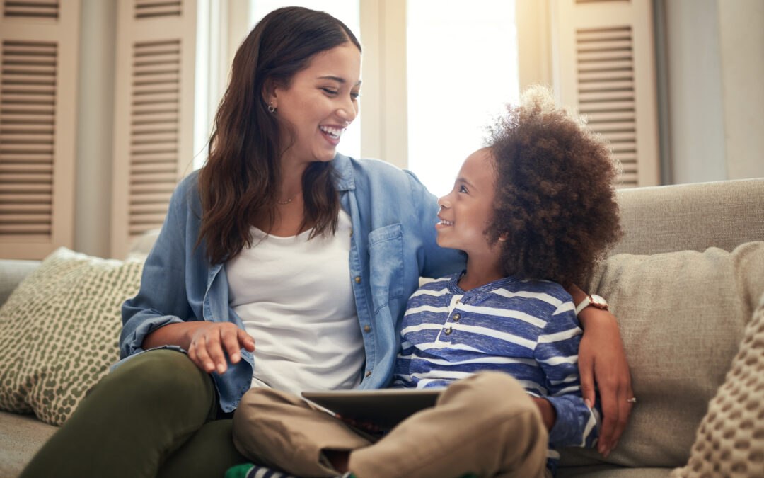 Mother and son sitting on couch smiling at each other with tablet on their laps representing positive relationship with technology
